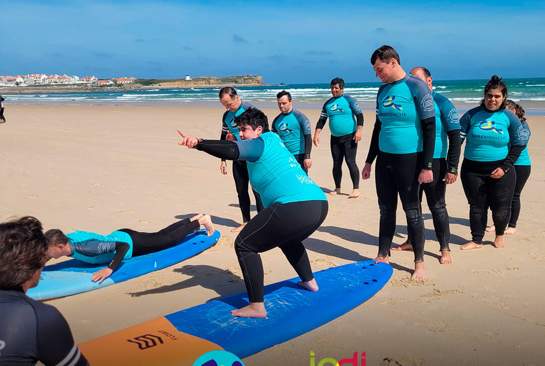 Pessoas apoiadas pela Cercipeniche na praia a realizar exercícios de aquecimento na prancha de surf antes de entrarem na água. Logo da Cercipeniche.