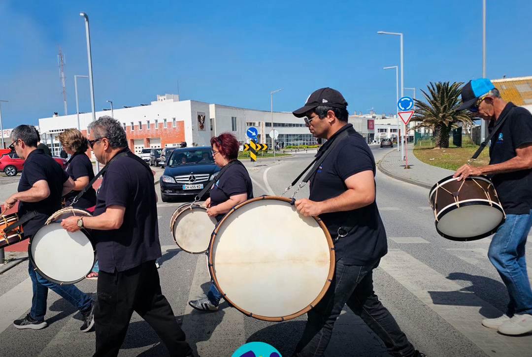 Grupo de pessoas apoiadas a tocar bombos na rua, durante o evento EnREDES. Logo da Cercipeniche.
