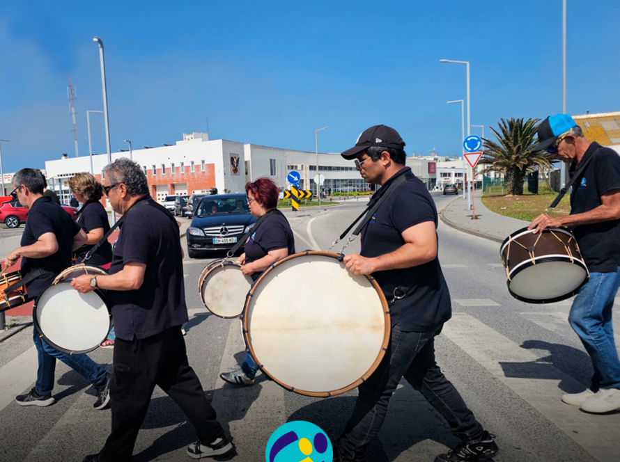 Grupo de pessoas apoiadas a tocar bombos na rua, durante o evento EnREDES. Logo da Cercipeniche.