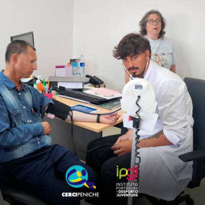 Médico a realizar a medição da tensão arterial a uma pessoa apoiada. Logo da Cercipeniche e do IPDJ.