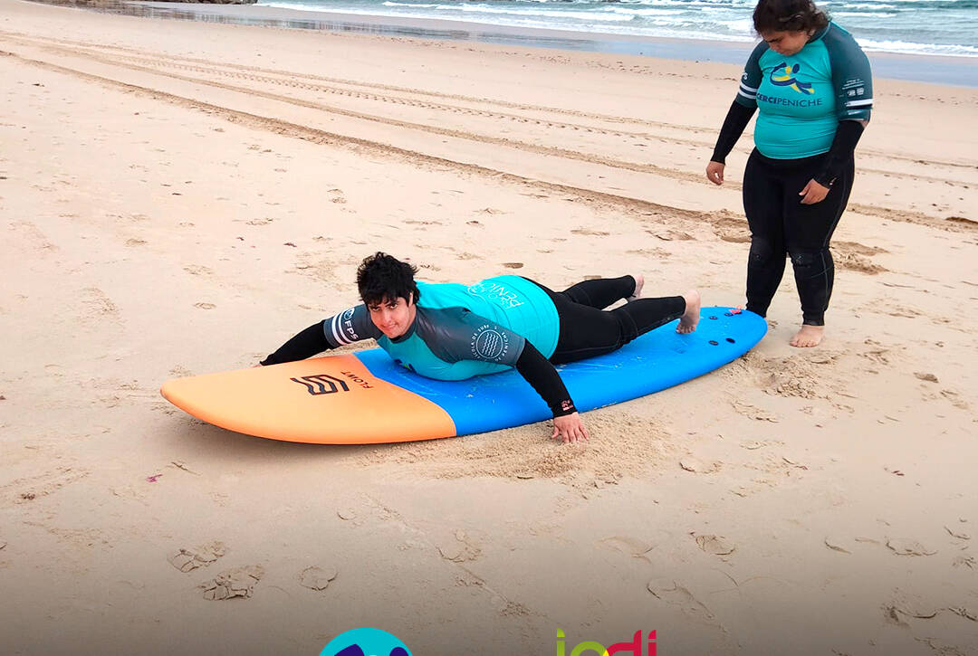 Pessoas apoiadas pela Cercipeniche em treino de equilíbrio sobre a prancha de surf, em aquecimento antes da prática no mar. Logo da Cercipeniche.