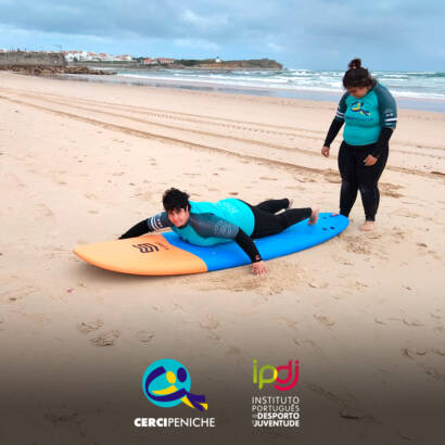 Pessoas apoiadas pela Cercipeniche em treino de equilíbrio sobre a prancha de surf, em aquecimento antes da prática no mar. Logo da Cercipeniche.