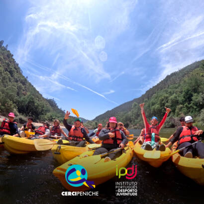 Grupo de participantes dentro de água, em caiaques, durante a descida do rio Mondego, cheios de alegria e entusiasmados pela experiência ao ar livre. Logo da Cercipeniche e do IPDJ.