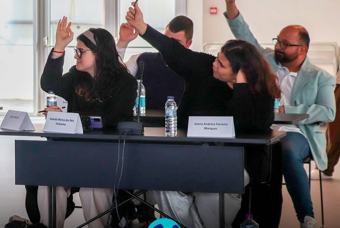 Vista geral de vários jovens participantes e adultos sentados em mesas dispostas em formato de reunião durante a Assembleia Municipal Jovem. Todos estão atentos ao decorrer da sessão. Sobre as mesas encontram-se microfones, garrafas de água e placas de identificação. Logótipo da Cercipeniche.