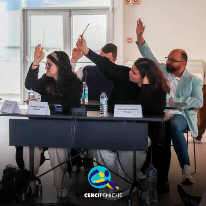 Vista geral de vários jovens participantes e adultos sentados em mesas dispostas em formato de reunião durante a Assembleia Municipal Jovem. Todos estão atentos ao decorrer da sessão. Sobre as mesas encontram-se microfones, garrafas de água e placas de identificação. Logótipo da Cercipeniche.