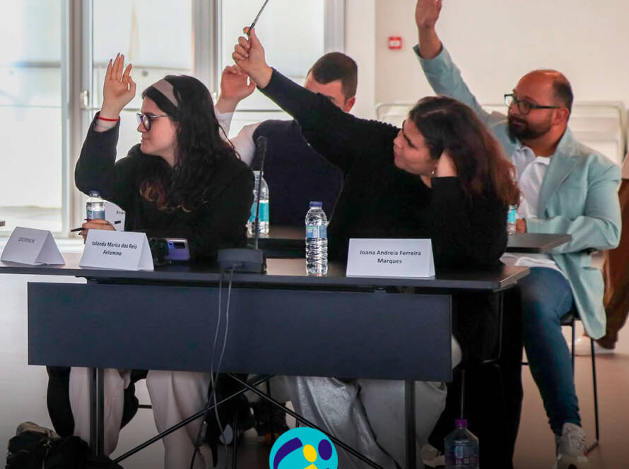 Vista geral de vários jovens participantes e adultos sentados em mesas dispostas em formato de reunião durante a Assembleia Municipal Jovem. Todos estão atentos ao decorrer da sessão. Sobre as mesas encontram-se microfones, garrafas de água e placas de identificação. Logótipo da Cercipeniche.