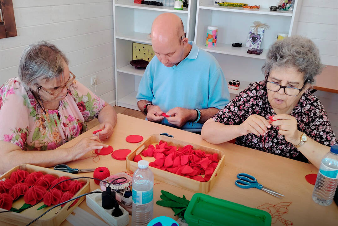 Foto com 3 pessoas sentadas à volta da mesa a coser manualmente peças de feltro vermelho para montar os cravos. Sobre a mesa encontram-se tabuleiros com materiais, tesouras, folhas em feltro verde, uma caixa de arrumação e uma garrafa de água. Logótipo da Cercipeniche.