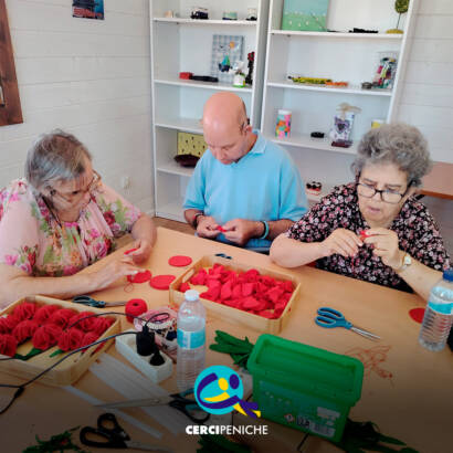 Foto com 3 pessoas sentadas à volta da mesa a coser manualmente peças de feltro vermelho para montar os cravos. Sobre a mesa encontram-se tabuleiros com materiais, tesouras, folhas em feltro verde, uma caixa de arrumação e uma garrafa de água. Logótipo da Cercipeniche.