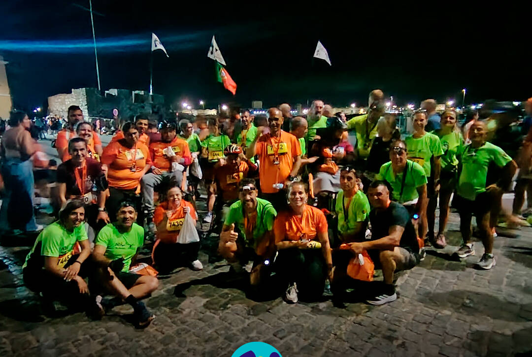 Grupo da Cercipeniche, acompanhado por voluntários da Equipa NEL, reúne-se no final da Corrida das Fogueiras para uma fotografia de grupo. Todos sorriem, exibindo as medalhas e celebrando a participação na prova. Logótipo da Cercipeniche.