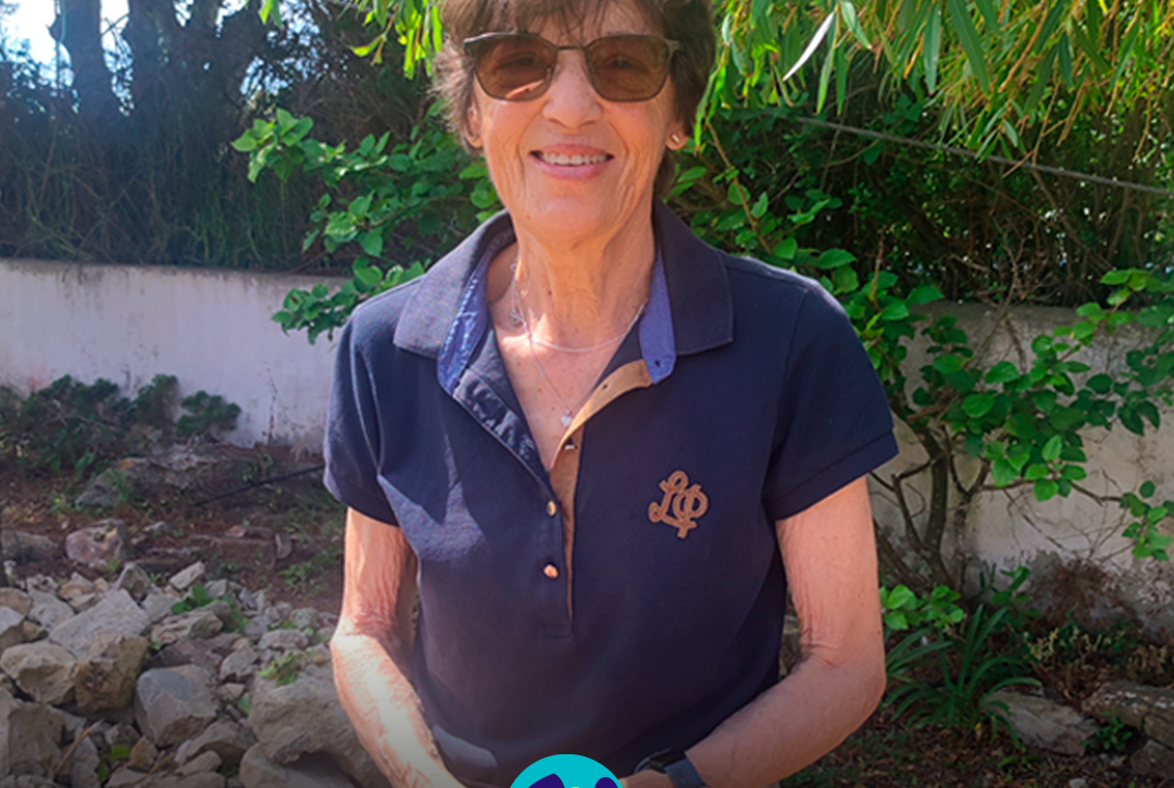 Foto da D. Antonieta, voluntária da Cercipeniche, a sorrir para a câmara. Está de pé, ao ar livre, com as mãos cruzadas à frente do corpo. Usa óculos de sol, uma camisola polo azul-escura de manga curta, umas calças claras e um relógio no pulso esquerdo. Ao fundo veem-se árvores, arbustos, pedras e um muro branco. Na parte inferior, o logótipo da Cercipeniche.