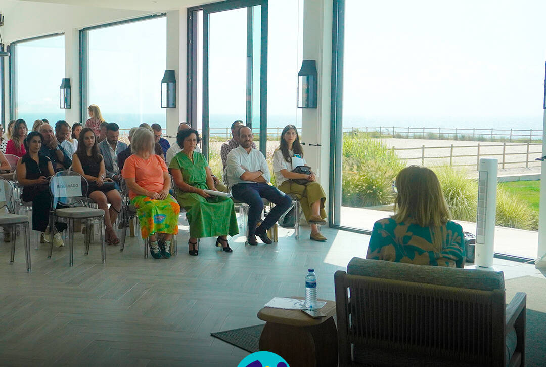 Vista geral da sala durante a sessão, com participantes sentados a assistir às intervenções. Ao fundo, grandes janelas deixam ver a paisagem exterior. Logo da Cercipeniche.