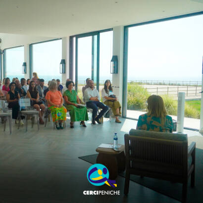 Vista geral da sala durante a sessão, com participantes sentados a assistir às intervenções. Ao fundo, grandes janelas deixam ver a paisagem exterior. Logo da Cercipeniche.
