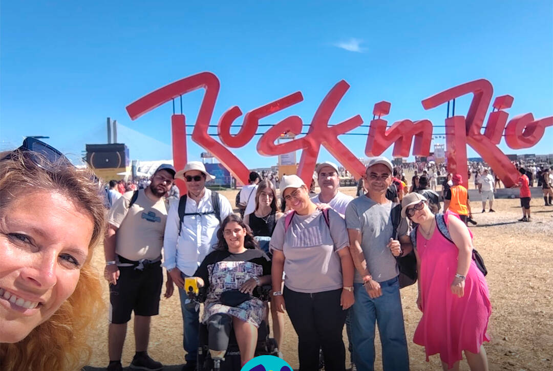 Pessoas apoiadas pela Cercipeniche pousam para a foto junto à entrada do Rock in Rio Lisboa. Ao fundo vê-se o logótipo do "Rock in Rio". Logótipo da Cercipeniche.