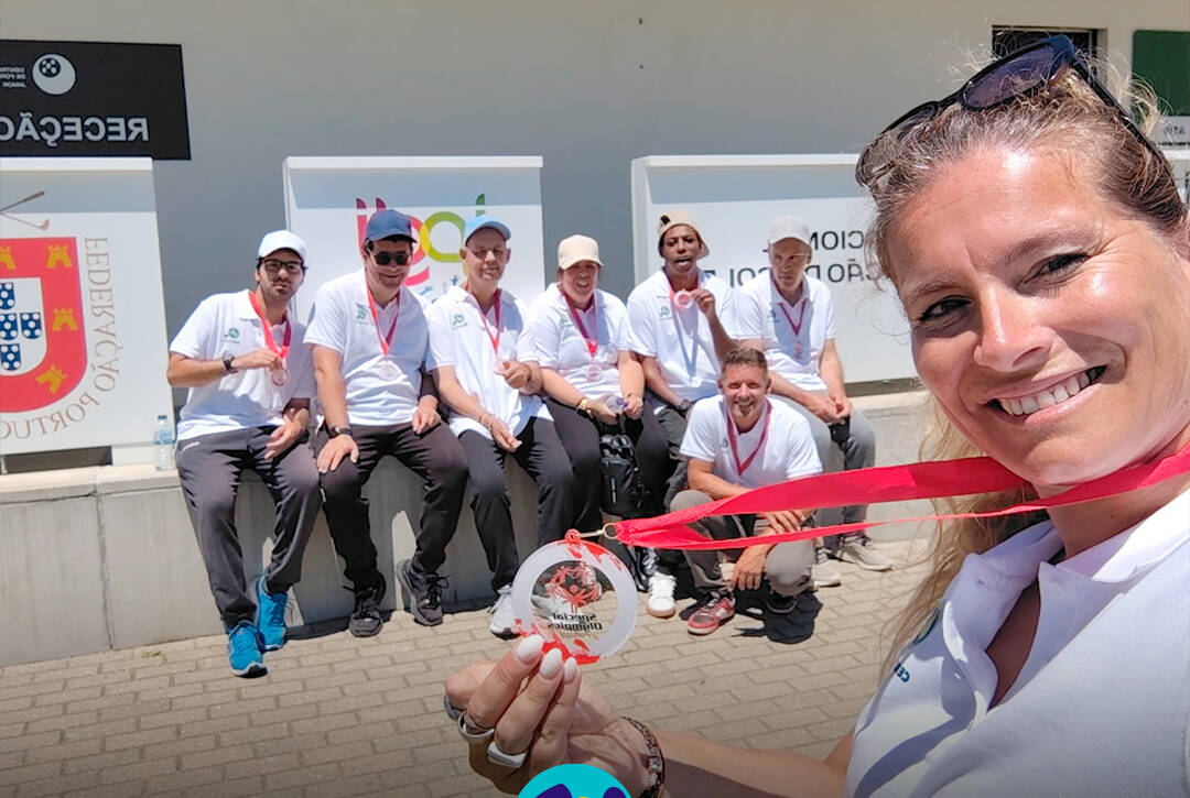 Em primeiro plano, uma pessoa tira uma selfie e segura uma medalha voltada para a câmara. Ao fundo, o grupo de pessoas apoiadas também com medalhas ao pescoço, está sentado em frente ao Centro Nacional de Formação de Golfe do Jamor. Logótipo da Cercipeniche.