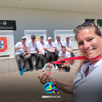 Em primeiro plano, uma pessoa tira uma selfie e segura uma medalha voltada para a câmara. Ao fundo, o grupo de pessoas apoiadas também com medalhas ao pescoço, está sentado em frente ao Centro Nacional de Formação de Golfe do Jamor. Logótipo da Cercipeniche.