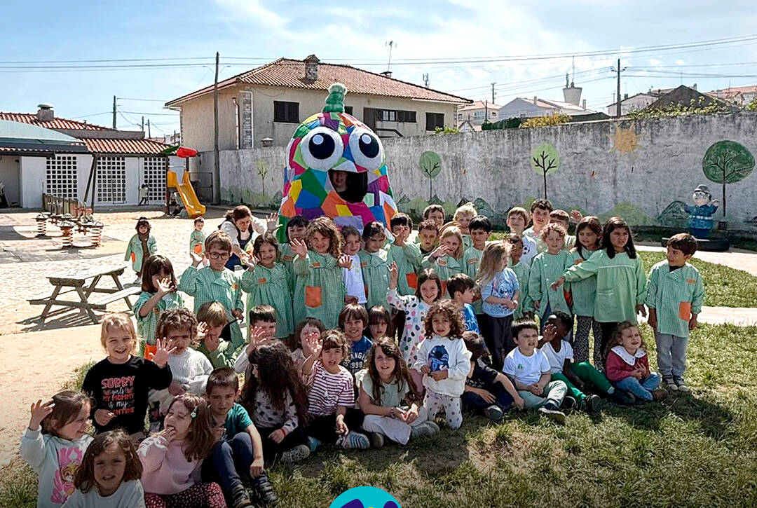 Foto de grupo, no pátio da Escola. Crianças sentadas no chão com a mascote do Pirilampo atrás. Logo da Cercipeniche.
