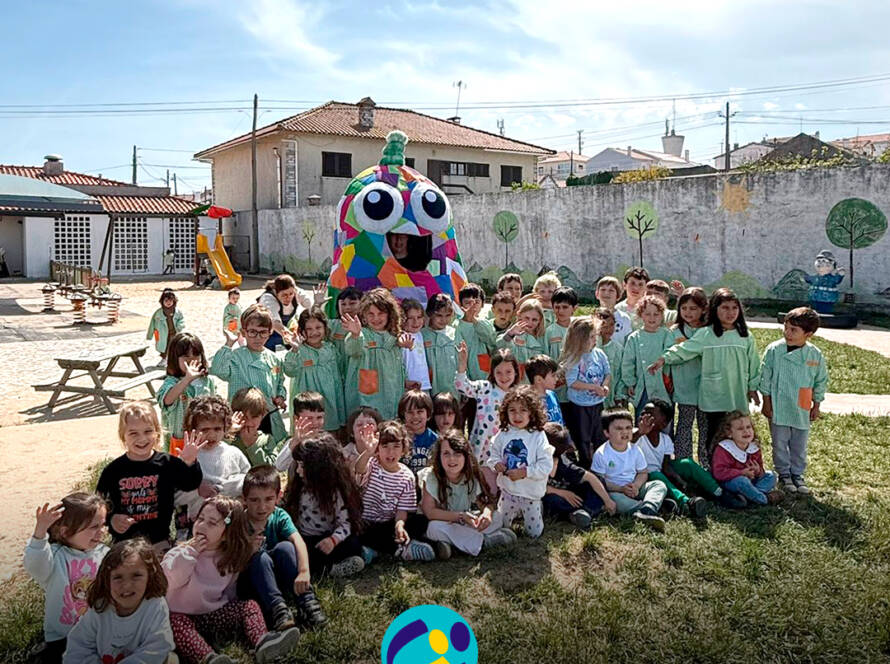 Foto de grupo, no pátio da Escola. Crianças sentadas no chão com a mascote do Pirilampo atrás. Logo da Cercipeniche.