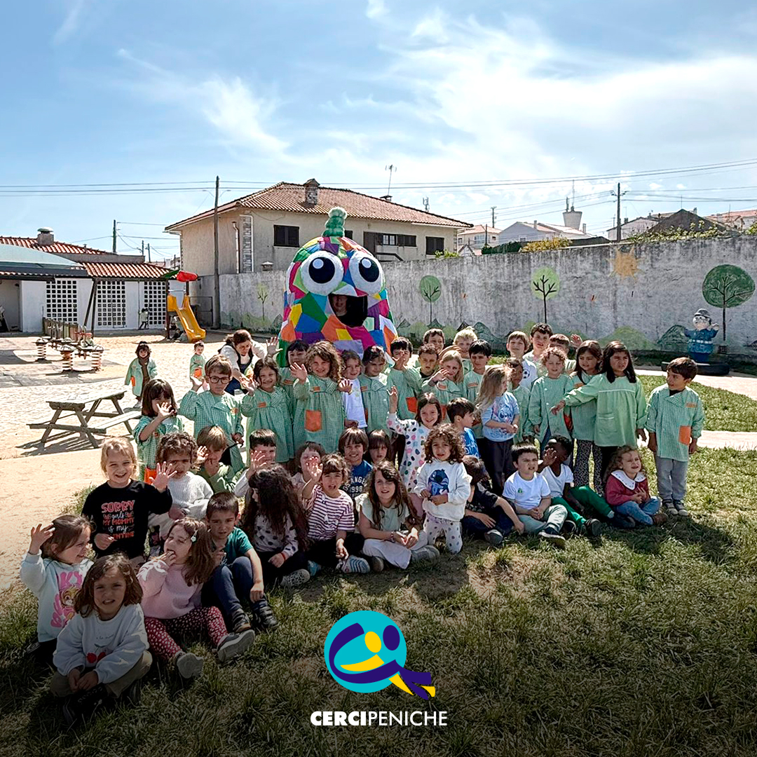 Foto de grupo, no pátio da Escola. Crianças sentadas no chão com a mascote do Pirilampo atrás. Logo da Cercipeniche.