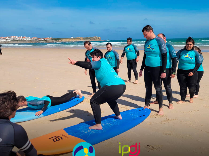 Pessoas apoiadas pela Cercipeniche na praia a realizar exercícios de aquecimento na prancha de surf antes de entrarem na água. Logo da Cercipeniche.