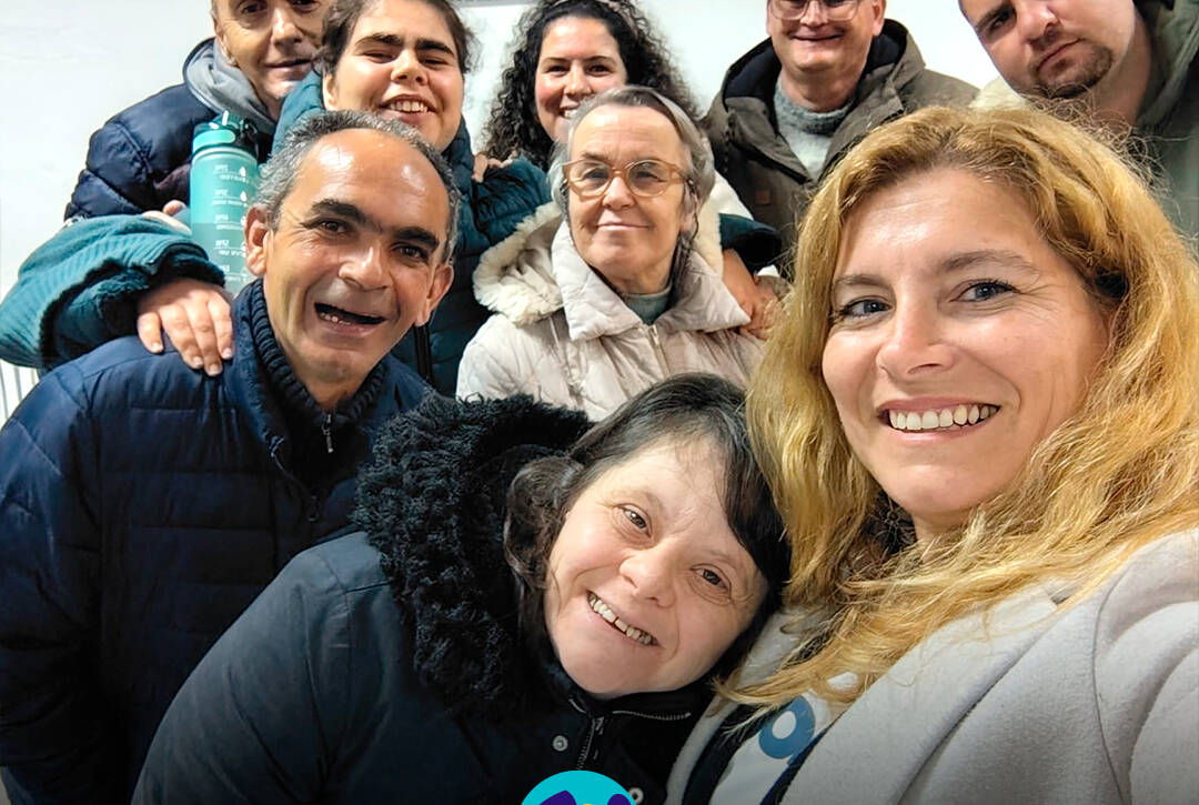 Grupo de pessoas apoiadas pela Cercipeniche posa com a animadora Raquel durante ensaio do rancho folclórico em Geraldes. Logo da Cercipeniche.