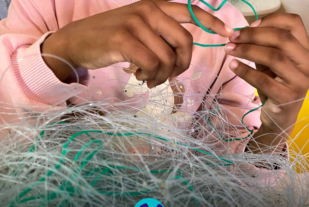 Mãos de uma criança a explorar redes de pesca no Atelier das Redes. Logo da Cercipeniche.