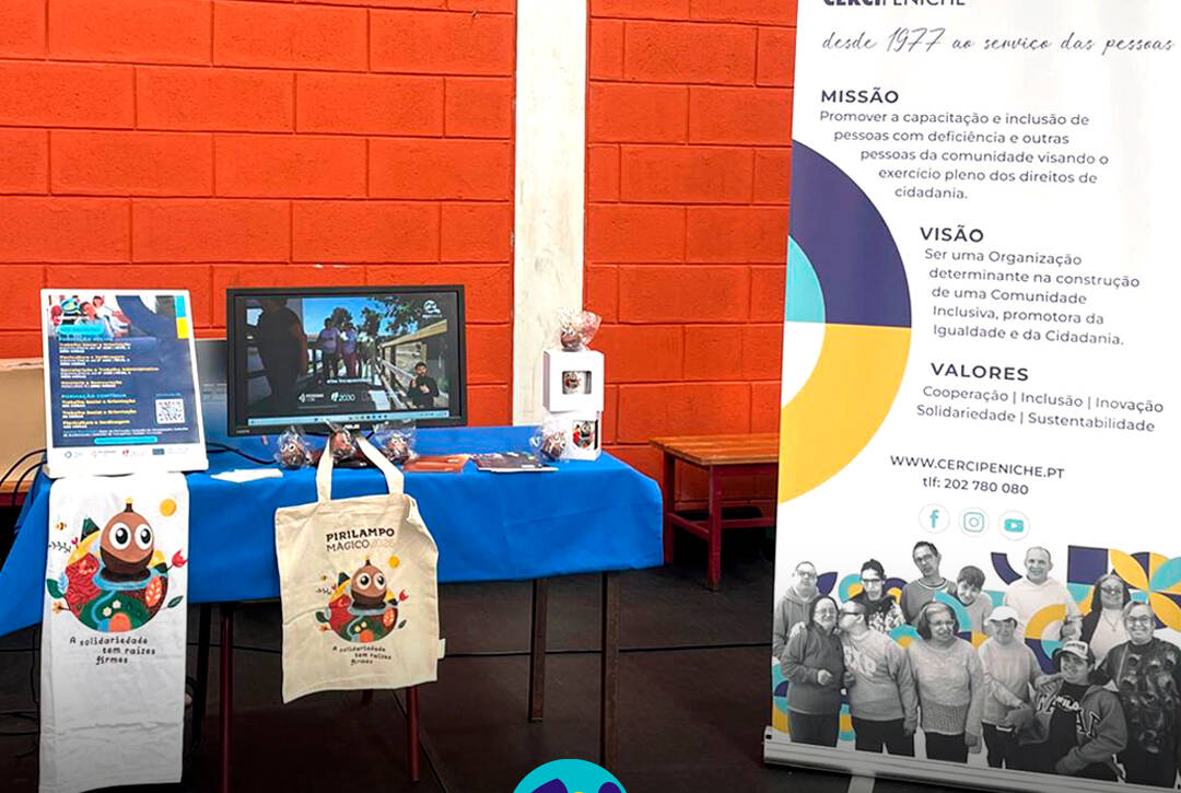 Mesa com materiais informativos da Cercipeniche - poster e flyers com as nossas formações, IRS solidário e evento Sunset Pirilampo Mágico, e produtos Pirilampo Mágico à venda. À direita, o nosso Roll Up indtitucional. Logo da Cercipeniche.