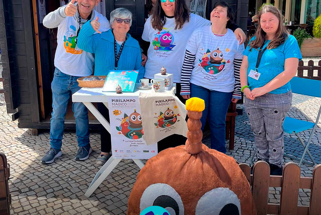 Pessoas apoiadas da Cercipeniche à entrada da Casinha do Pirilampo Mágico, preparadas para iniciar a manhã de vendas. Na foto vê-se produtos expostos numa mesa e a mascote do Pirilampo Mágico. Logo da Cercipeniche.