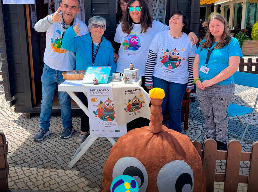 Pessoas apoiadas da Cercipeniche à entrada da Casinha do Pirilampo Mágico, preparadas para iniciar a manhã de vendas. Na foto vê-se produtos expostos numa mesa e a mascote do Pirilampo Mágico. Logo da Cercipeniche.