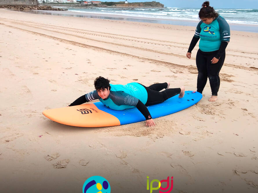 Pessoas apoiadas pela Cercipeniche em treino de equilíbrio sobre a prancha de surf, em aquecimento antes da prática no mar. Logo da Cercipeniche.