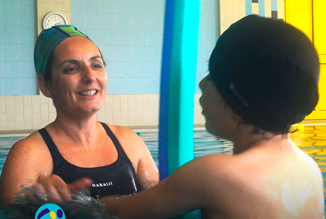 CRI – Centro de Recursos para a Inclusão: Técnica da Cercipeniche sorri enquanto acompanha um menino numa atividade realizada dentro da piscina. Logo da Cercipeniche.
