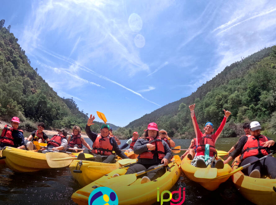 Grupo de participantes dentro de água, em caiaques, durante a descida do rio Mondego, cheios de alegria e entusiasmados pela experiência ao ar livre. Logo da Cercipeniche e do IPDJ.