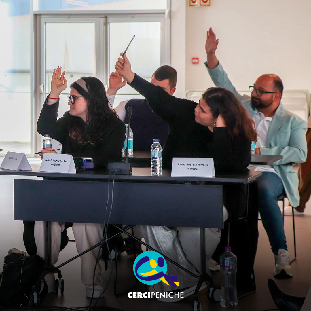 Vista geral de vários jovens participantes e adultos sentados em mesas dispostas em formato de reunião durante a Assembleia Municipal Jovem. Todos estão atentos ao decorrer da sessão. Sobre as mesas encontram-se microfones, garrafas de água e placas de identificação. Logótipo da Cercipeniche.