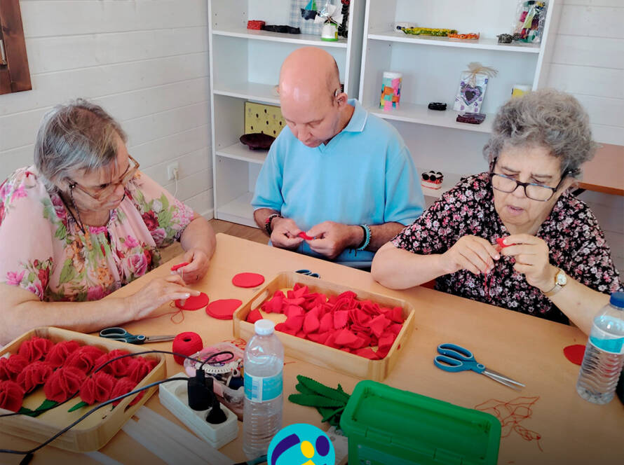 Foto com 3 pessoas sentadas à volta da mesa a coser manualmente peças de feltro vermelho para montar os cravos. Sobre a mesa encontram-se tabuleiros com materiais, tesouras, folhas em feltro verde, uma caixa de arrumação e uma garrafa de água. Logótipo da Cercipeniche.