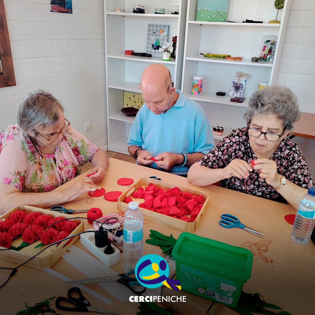 Foto com 3 pessoas sentadas à volta da mesa a coser manualmente peças de feltro vermelho para montar os cravos. Sobre a mesa encontram-se tabuleiros com materiais, tesouras, folhas em feltro verde, uma caixa de arrumação e uma garrafa de água. Logótipo da Cercipeniche.