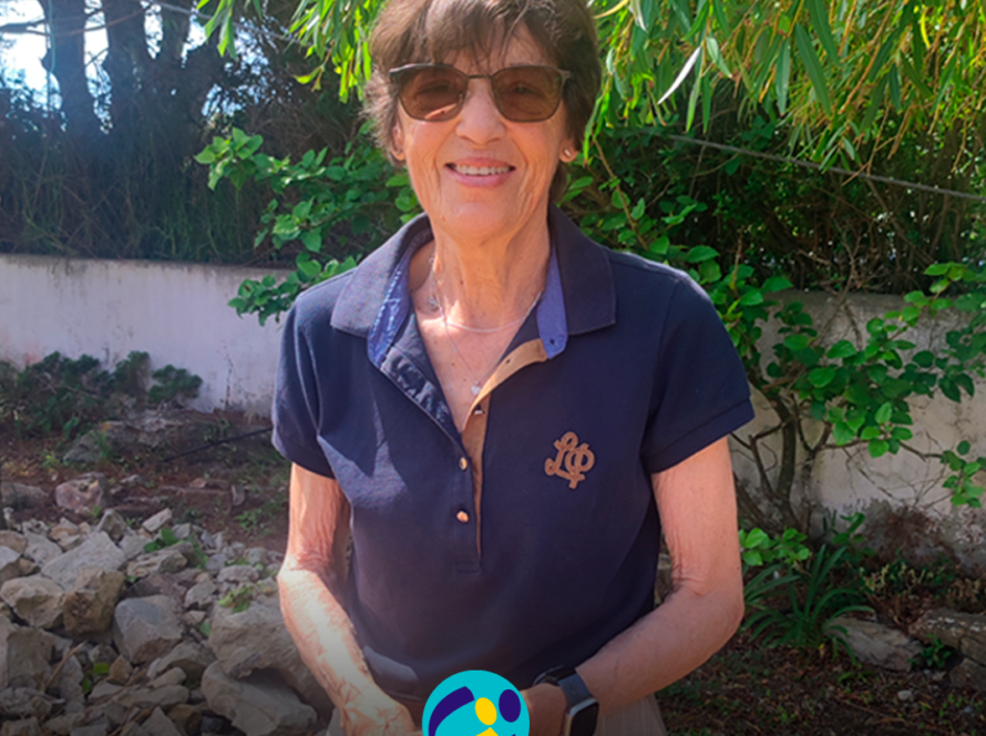 Foto da D. Antonieta, voluntária da Cercipeniche, a sorrir para a câmara. Está de pé, ao ar livre, com as mãos cruzadas à frente do corpo. Usa óculos de sol, uma camisola polo azul-escura de manga curta, umas calças claras e um relógio no pulso esquerdo. Ao fundo veem-se árvores, arbustos, pedras e um muro branco. Na parte inferior, o logótipo da Cercipeniche.