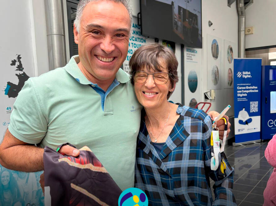 Sérgio Leandro e a D. Antonieta posam abraçados, a sorrir para a fotografia, enquanto ele segura um saco produzido pela Cercipeniche. Logo da Cercipeniche.