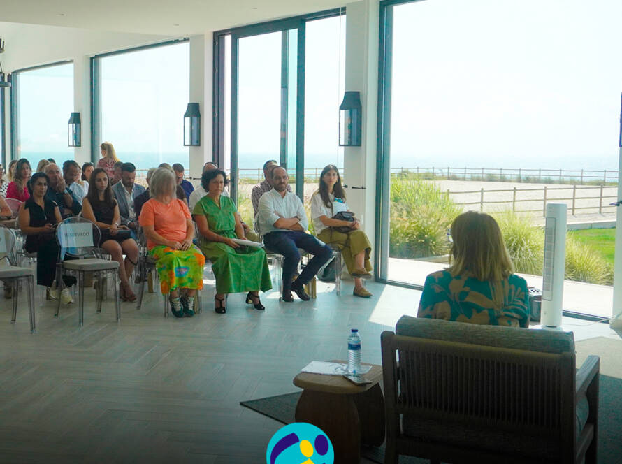 Vista geral da sala durante a sessão, com participantes sentados a assistir às intervenções. Ao fundo, grandes janelas deixam ver a paisagem exterior. Logo da Cercipeniche.