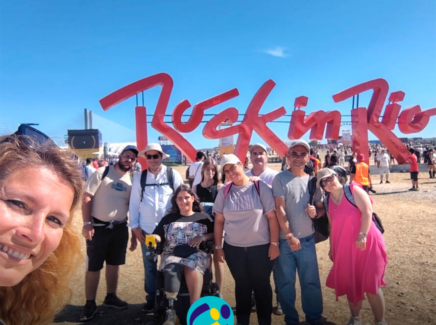 Pessoas apoiadas pela Cercipeniche pousam para a foto junto à entrada do Rock in Rio Lisboa. Ao fundo vê-se o logótipo do "Rock in Rio". Logótipo da Cercipeniche.