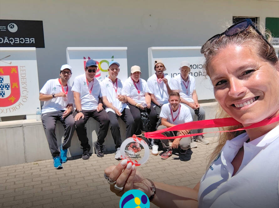 Em primeiro plano, uma pessoa tira uma selfie e segura uma medalha voltada para a câmara. Ao fundo, o grupo de pessoas apoiadas também com medalhas ao pescoço, está sentado em frente ao Centro Nacional de Formação de Golfe do Jamor. Logótipo da Cercipeniche.