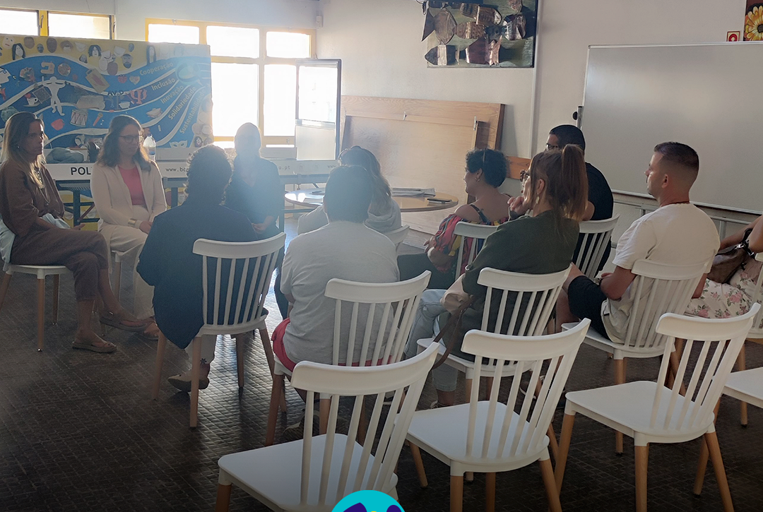 Fotografia de uma sessão de (in)formação realizada numa sala da CERCIPENICHE. Um grupo de pessoas está sentado em círculo, em cadeiras brancas, a conversar. Ao fundo é visível um painel colorido com palavras como “cooperação”, “inclusão”, “solidariedade” e “sustentabilidade”. Na parte inferior da imagem encontra-se o logótipo da CERCIPENICHE.