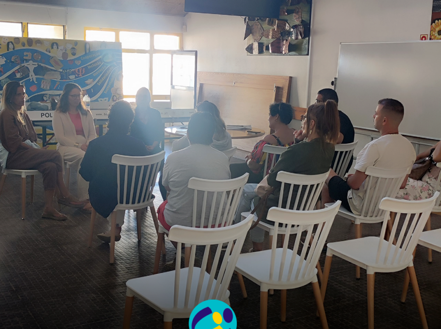 Fotografia de uma sessão de (in)formação realizada numa sala da CERCIPENICHE. Um grupo de pessoas está sentado em círculo, em cadeiras brancas, a conversar. Ao fundo é visível um painel colorido com palavras como “cooperação”, “inclusão”, “solidariedade” e “sustentabilidade”. Na parte inferior da imagem encontra-se o logótipo da CERCIPENICHE.