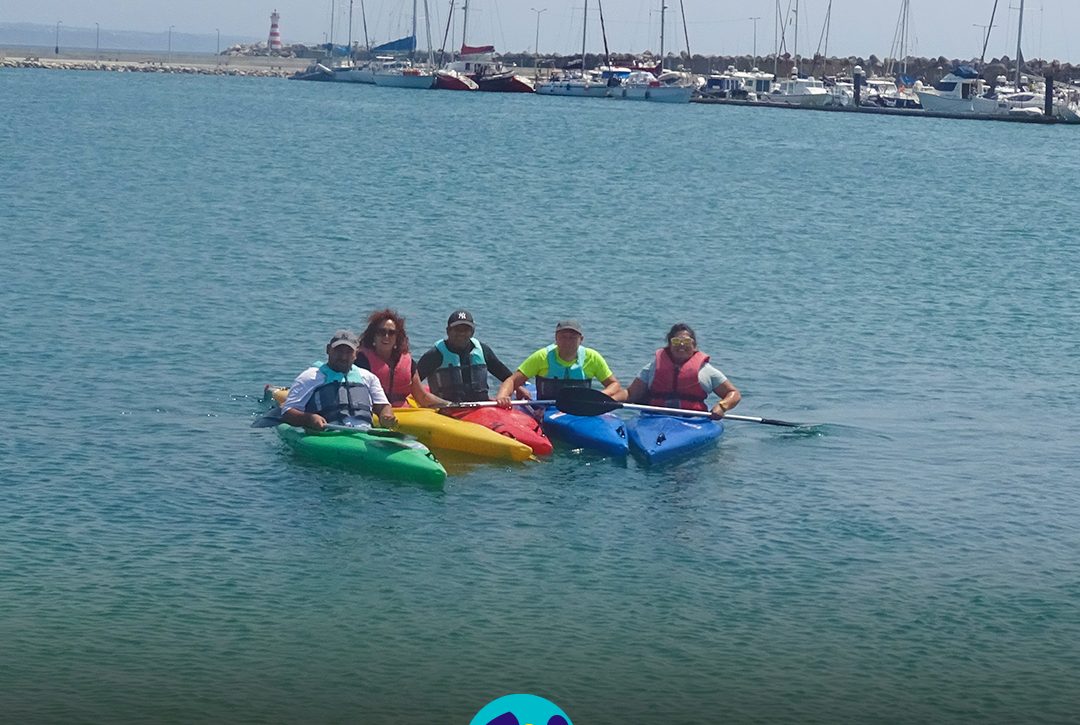 Pessoas apoiadas pela Cercipeniche e monitoras estão em caiaques, juntas na água, durante uma atividade de canoagem em Peniche. Os caiaques são verdes, amarelo, vermelho e azul. Todas as pessoas usam coletes de proteção e seguram remos. Ao fundo encontra-se a marina, com vários barcos à vela e outras embarcações. Mais à esquerda é visível um farol com riscas vermelhas e brancas, junto ao molhe. Na parte inferior da imagem aparece o logótipo da Cercipeniche.