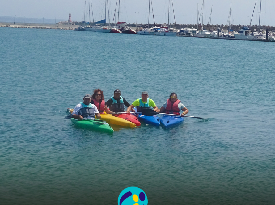 Pessoas apoiadas pela Cercipeniche e monitoras estão em caiaques, juntas na água, durante uma atividade de canoagem em Peniche. Os caiaques são verdes, amarelo, vermelho e azul. Todas as pessoas usam coletes de proteção e seguram remos. Ao fundo encontra-se a marina, com vários barcos à vela e outras embarcações. Mais à esquerda é visível um farol com riscas vermelhas e brancas, junto ao molhe. Na parte inferior da imagem aparece o logótipo da Cercipeniche.