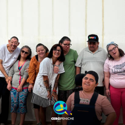 Fotografia de grupo, em ambiente interior, com 8 pessoas. Ao fundo, 7 pessoas estão lado a lado, a sorrir e a olhar para a câmara. Da esquerda para a direita, vê-se uma pessoa apoiada pela Cercipeniche de camisola branca, outra de camisola cinzenta e calças estampadas, outra de camisola laranja, uma pessoa de camisola branca, outra de camisola verde, outra de camisola cinzenta e boné preto e uma pessoa de camisola rosa e calças vermelhas. Em primeiro plano, está uma pessoa apoiada sentada numa cadeira de rodas, com uma camisola rosa e um suporte preto. Ao fundo, vê-se uma parede clara. Logótipo da Cercipeniche.