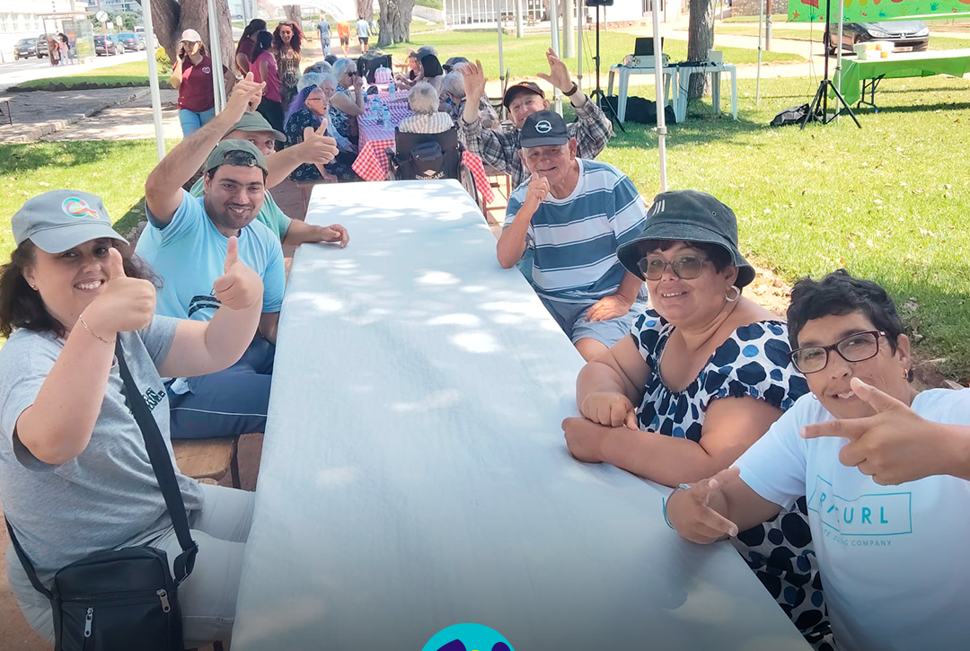 Várias pessoas apoiadas pela Cercipeniche sentadas à volta de uma mesa comprida, no exterior, durante a celebração do Dia dos Avós. Ao fundo estão outras pessoas sentadas em mesas, debaixo de tendas azuis. Do lado direito vê-se um painel verde com as palavras “Dia dos Avós”. Na parte inferior da imagem aparece o logótipo da Cercipeniche.