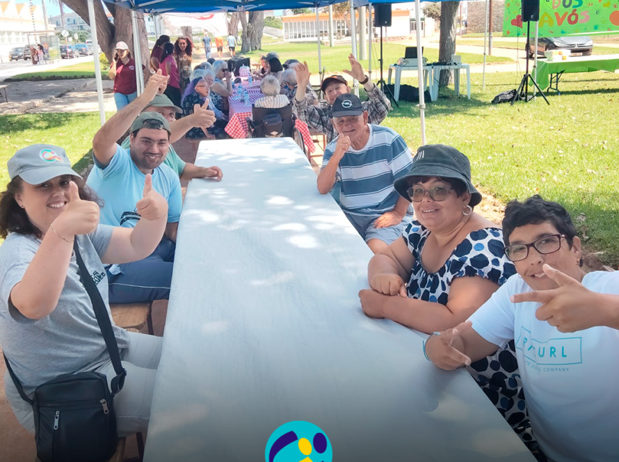Várias pessoas apoiadas pela Cercipeniche sentadas à volta de uma mesa comprida, no exterior, durante a celebração do Dia dos Avós. Ao fundo estão outras pessoas sentadas em mesas, debaixo de tendas azuis. Do lado direito vê-se um painel verde com as palavras “Dia dos Avós”. Na parte inferior da imagem aparece o logótipo da Cercipeniche.