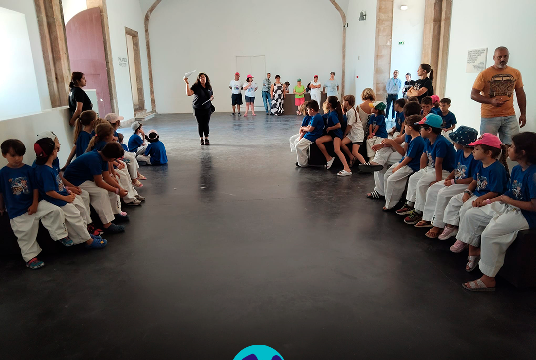 Interior do Museu Nacional Resistência e Liberdade – Fortaleza de Peniche. Várias crianças, vestidas com camisolas azuis e calças brancas, estão sentadas em 2 filas, uma de cada lado de uma sala ampla. No centro, a monitora está de pé enquanto fala para o grupo. Ao fundo encontram-se várias pessoas apoiadas pela Cercipeniche e outros adultos. A sala tem paredes brancas, chão escuro e portas e arcos de pedra. Na parte inferior da imagem aparece o logótipo da Cercipeniche.