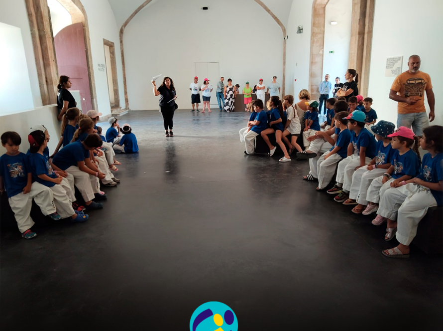 Interior do Museu Nacional Resistência e Liberdade – Fortaleza de Peniche. Várias crianças, vestidas com camisolas azuis e calças brancas, estão sentadas em 2 filas, uma de cada lado de uma sala ampla. No centro, a monitora está de pé enquanto fala para o grupo. Ao fundo encontram-se várias pessoas apoiadas pela Cercipeniche e outros adultos. A sala tem paredes brancas, chão escuro e portas e arcos de pedra. Na parte inferior da imagem aparece o logótipo da Cercipeniche.