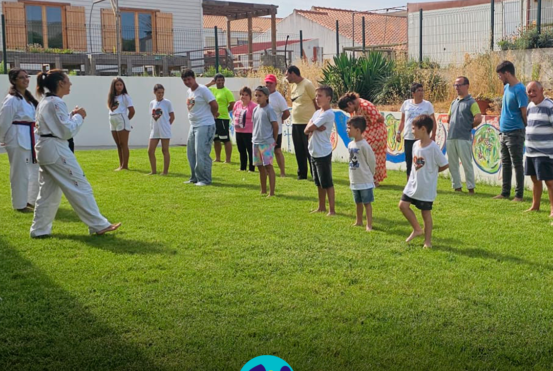 Num espaço exterior relvado da Cercipeniche, um grupo de pessoas apoiadas participa numa atividade de Taekwondo, acompanhando os movimentos demonstrados por duas instrutoras. Logo da Cercipeniche.