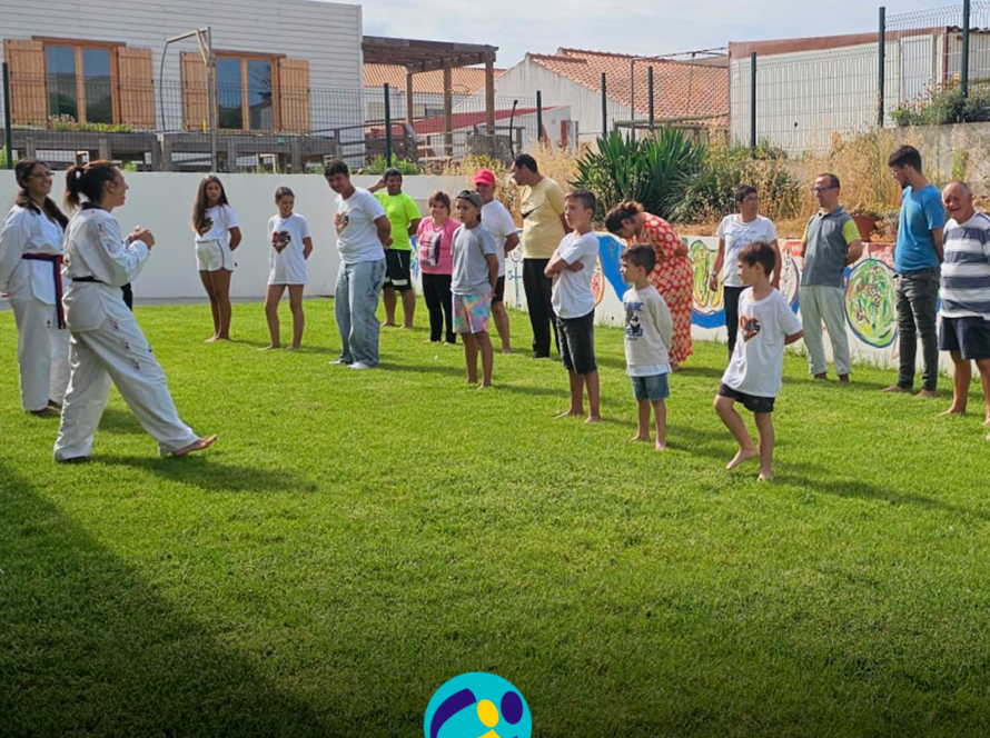 Num espaço exterior relvado da Cercipeniche, um grupo de pessoas apoiadas participa numa atividade de Taekwondo, acompanhando os movimentos demonstrados por duas instrutoras. Logo da Cercipeniche.